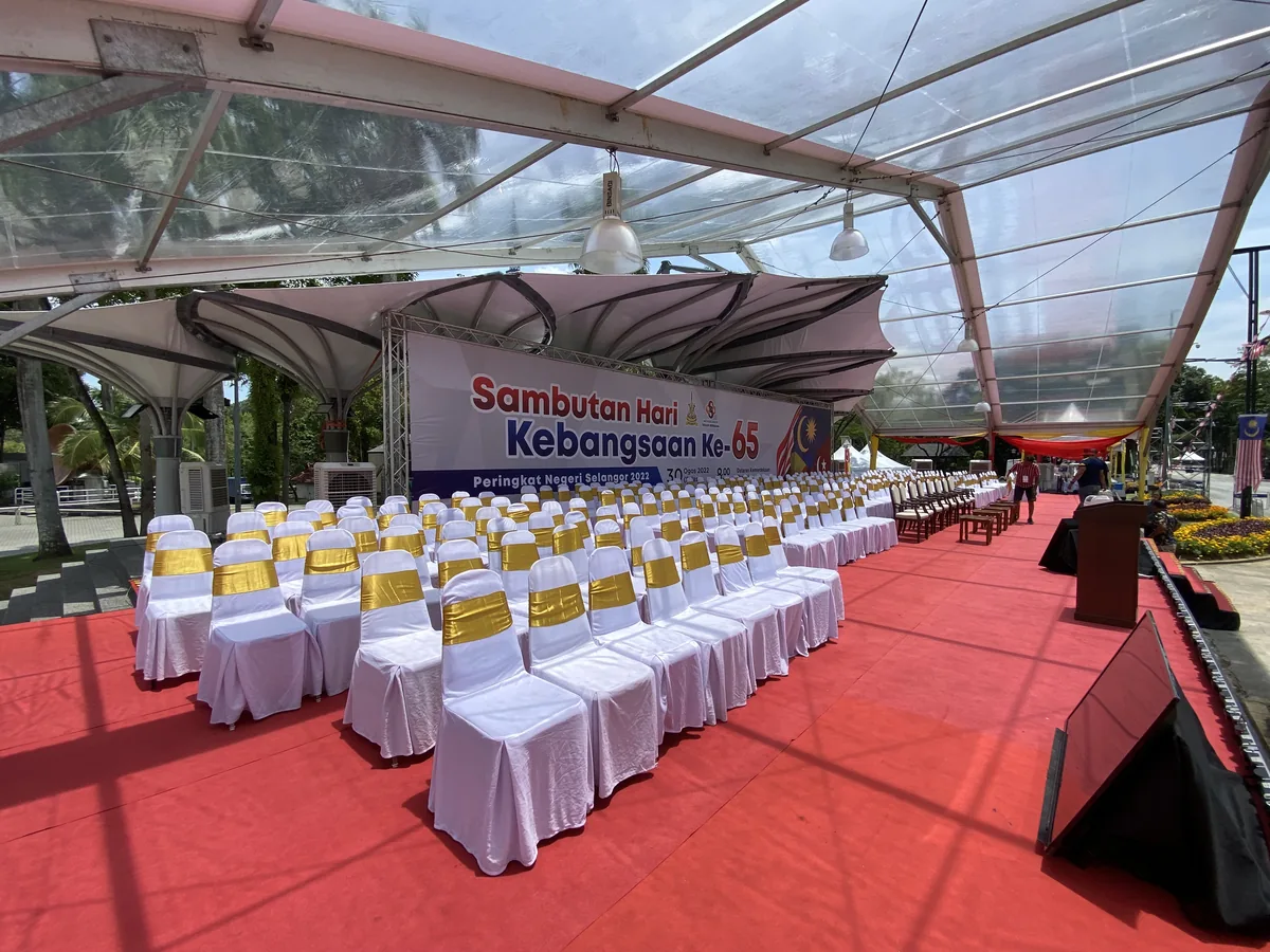 Rows of sashed chairs on red carpet under a clear canopy facing a stage banner