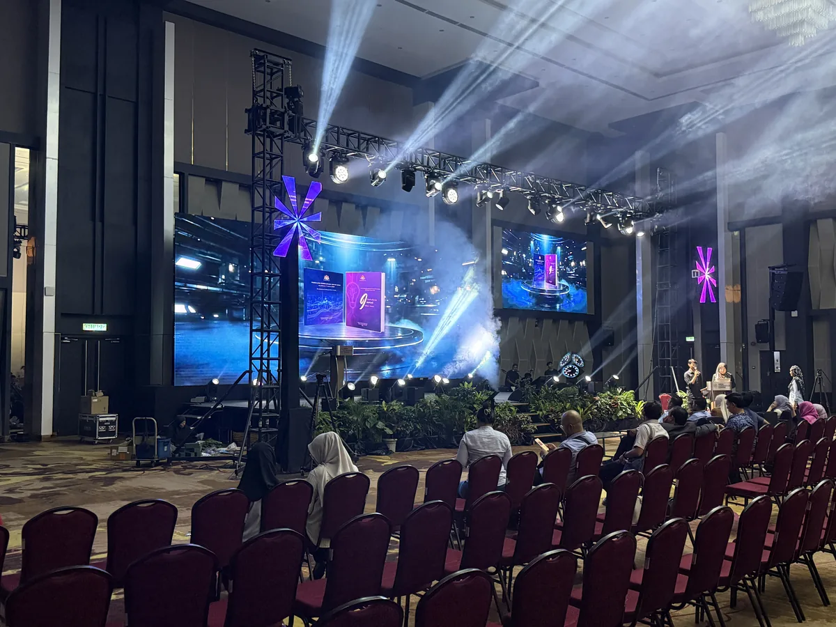 Conference stage with LED screens, a lighting tower and beams above rows of chairs