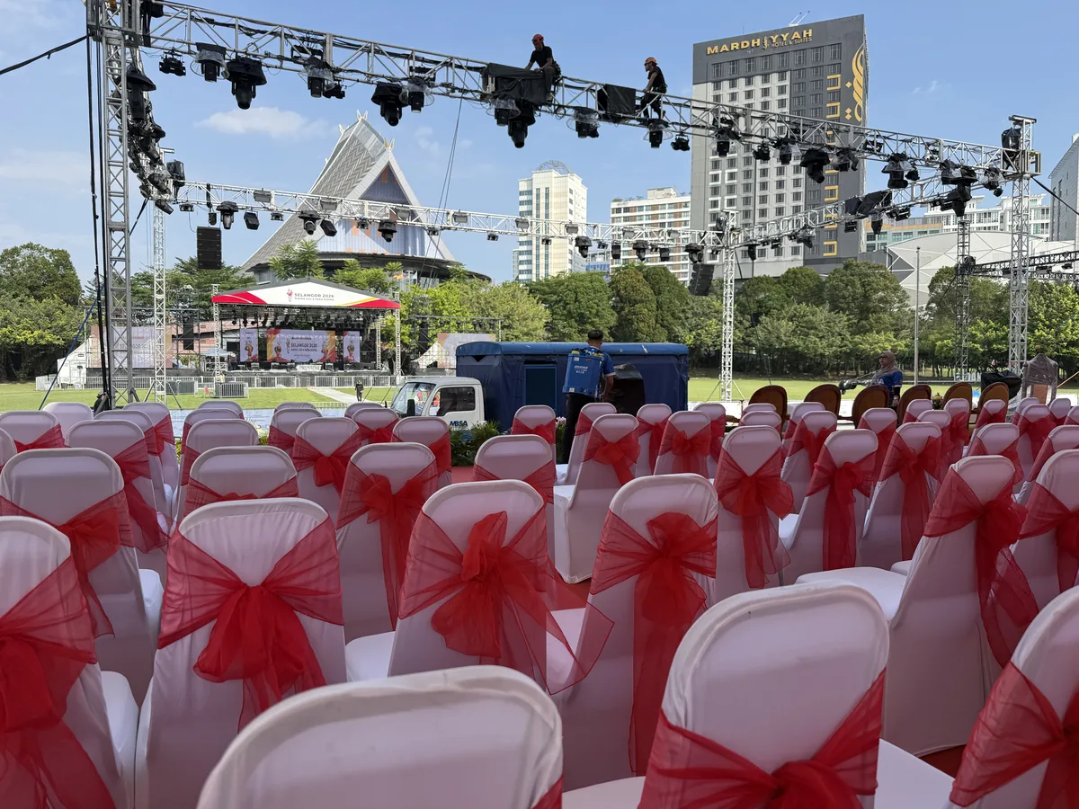 Rows of sashed chairs facing an outdoor stage while crew work on the lighting truss